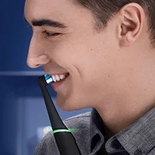 A man smiles while brushing his teeth with a black electric toothbrush that has a green light ring.