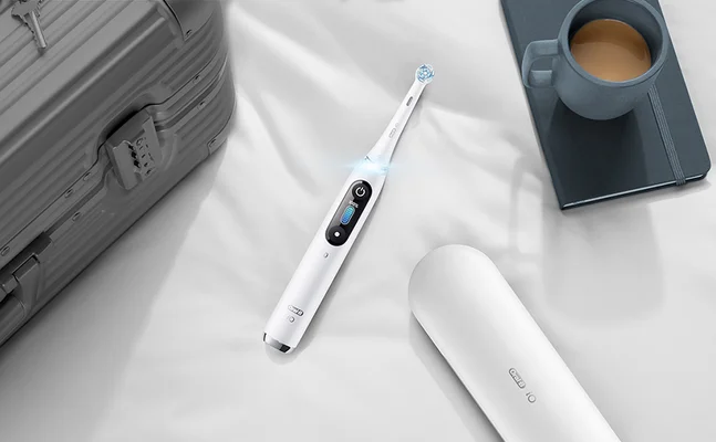 Oral-B iO elektrische Zahnbürste, zusammen mit Reiseetui, Koffer und Kaffeetasse auf einem Bettlaken.