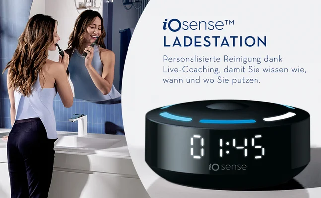 A woman brushes her teeth in a bathroom reflected in a mirror next to a black iOsense Ladestation device displaying 0 1:45 and iO sense, with overlay text reading iOsense™ LADESTATION Personalisierte Reinigung dank Live-Coaching, damit Sie wissen wie, wann und wo Sie putzen.