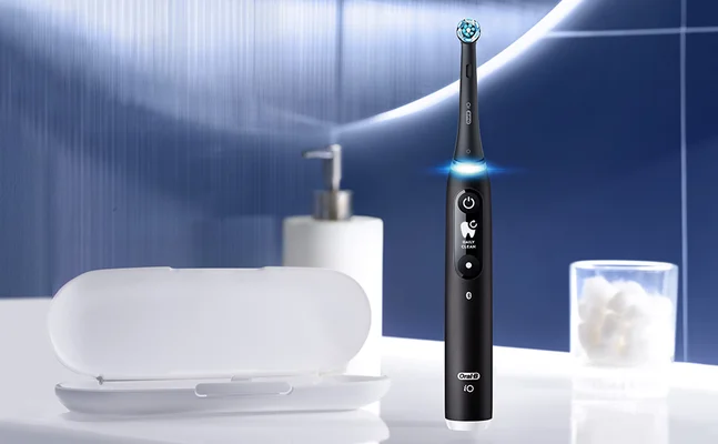 A black Oral-B iO electric toothbrush with text Oral-B iO and DAILY CLEAN stands next to a white travel case, soap dispenser, and cotton balls in a bathroom setting.