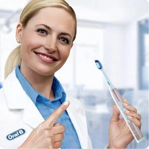 Frau im weißen Kittel zeigt auf eine Oral-B Zahnbürste und lächelt.
